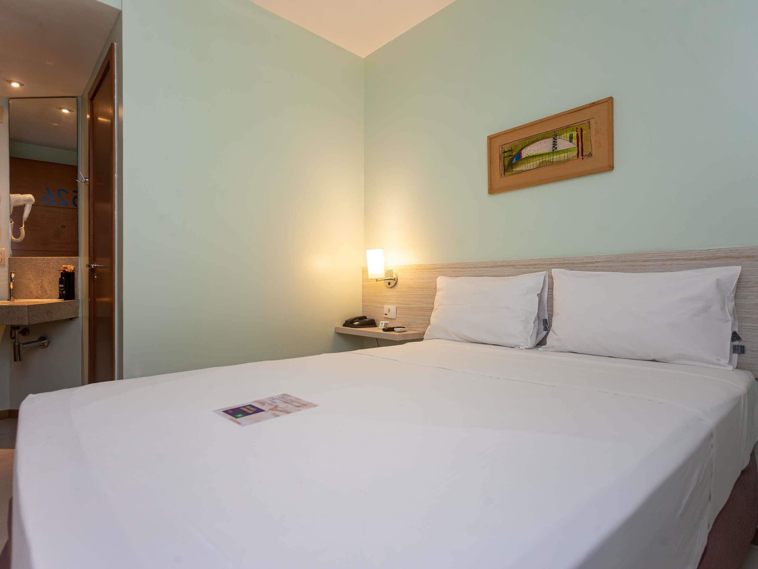 Quarto de hotel com cama, mesa de cabeceira, luminária na parede e porta de acesso a um banheiro.