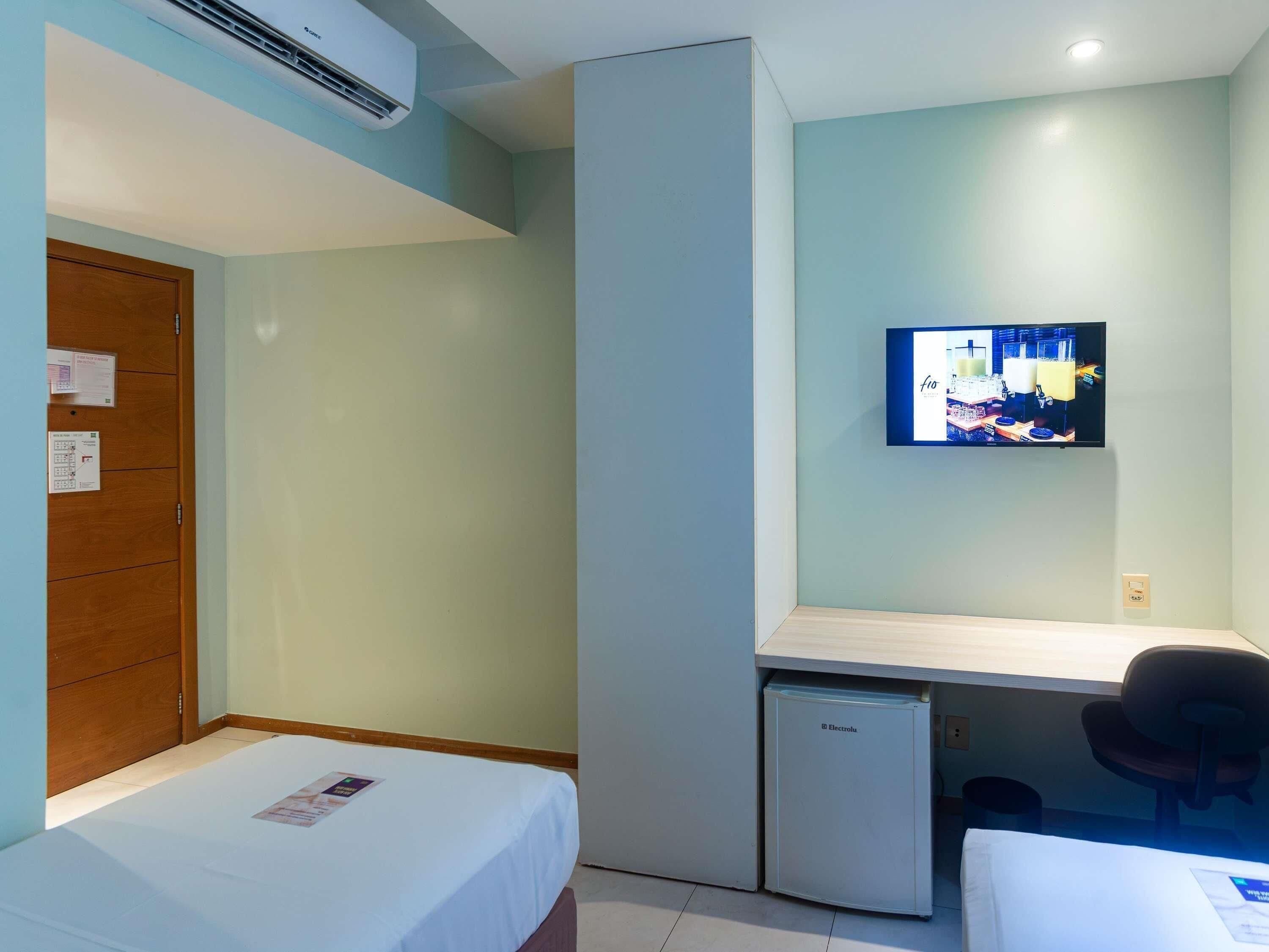 Habitación de hotel con cama, escritorio con silla, refrigerador pequeño y televisor montado en la pared.