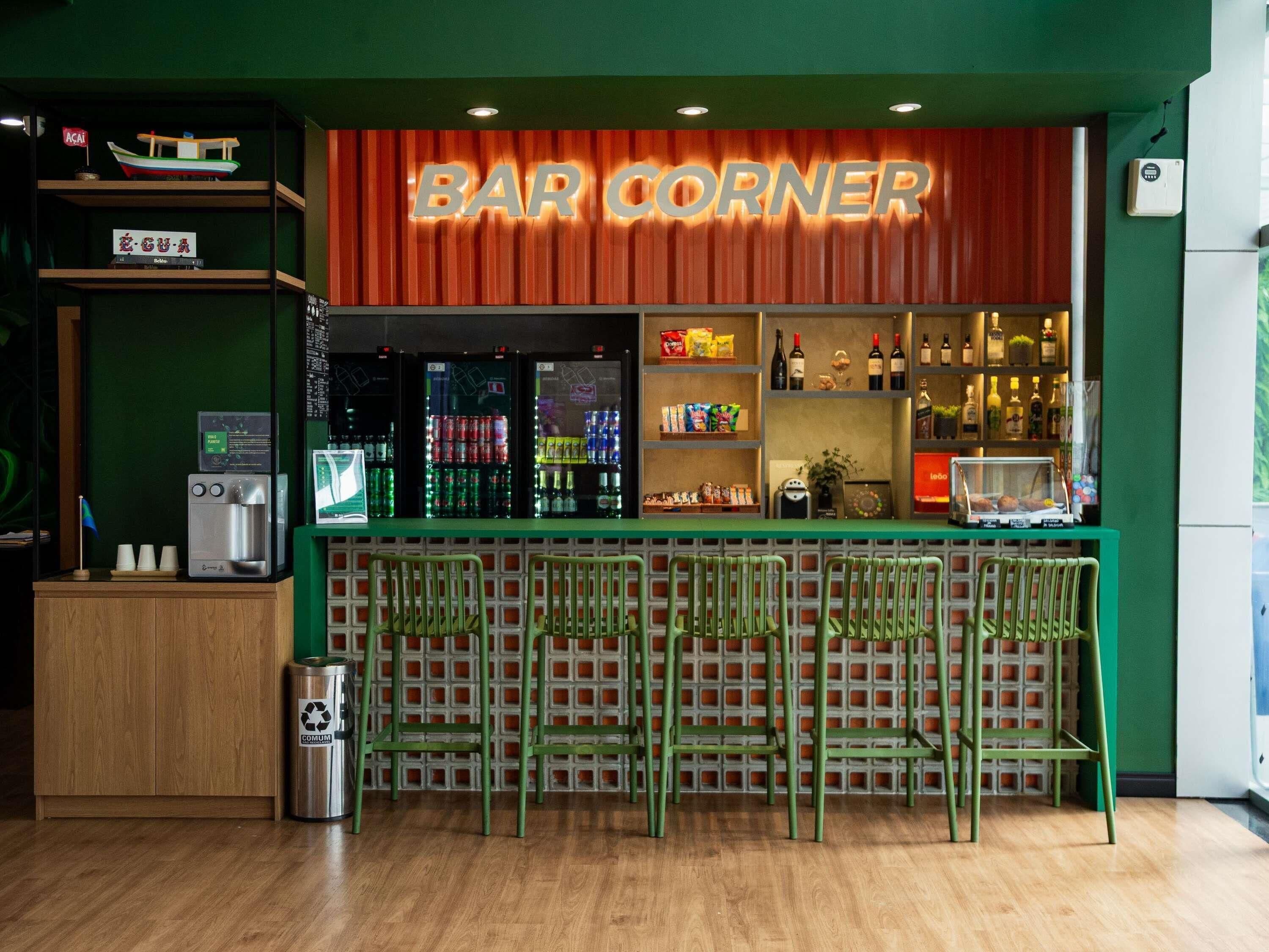 Uma área de bar com um letreiro indicando 'BAR CORNER', prateleiras com lanches e bebidas, e um bebedouro.