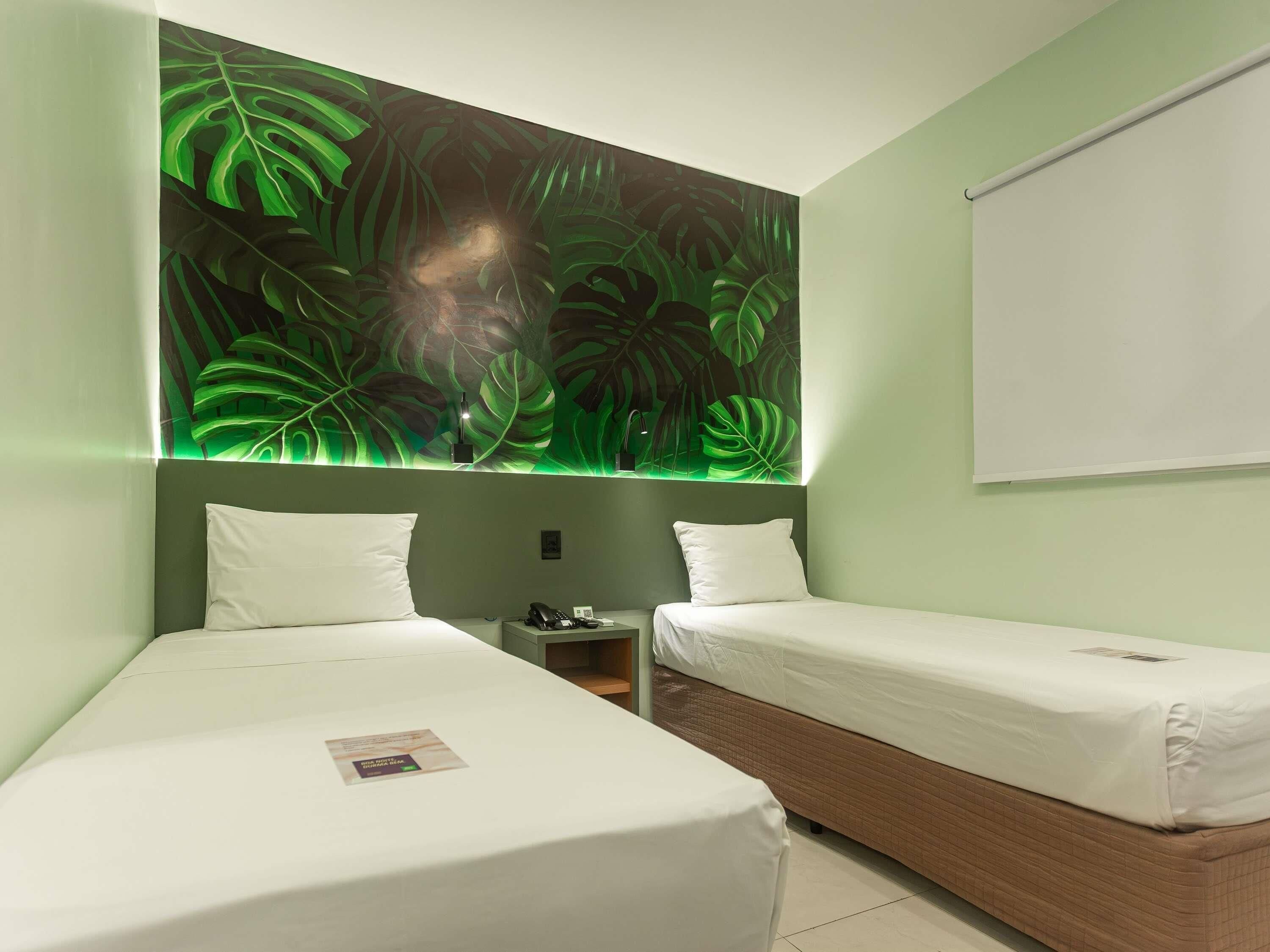 A hotel room with two beds, a nightstand, and a large wall mural of tropical leaves.