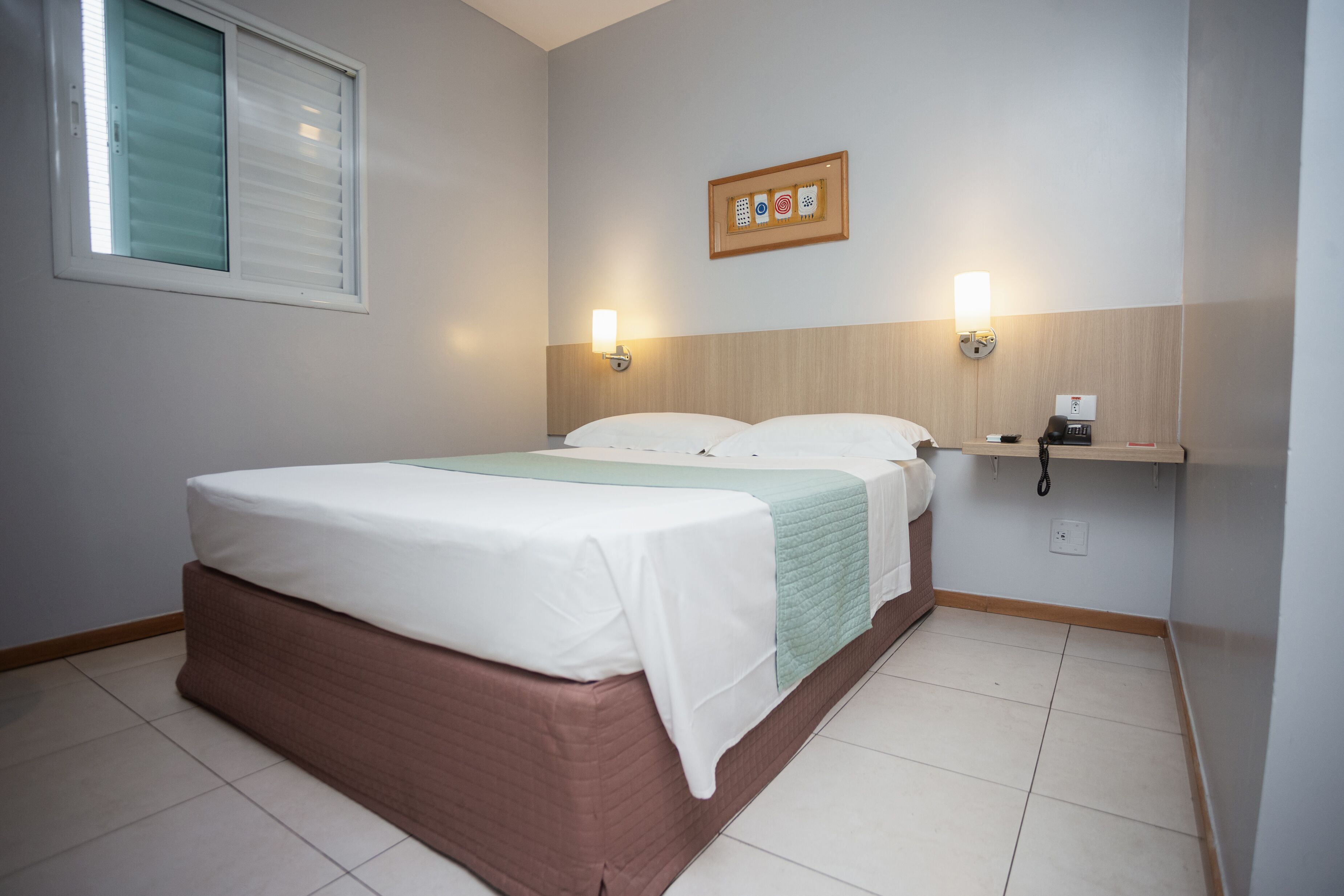 Une chambre d’hôtel comprenant un lit, deux tables de chevet, une étagère fixée au mur et une fenêtre avec des stores.