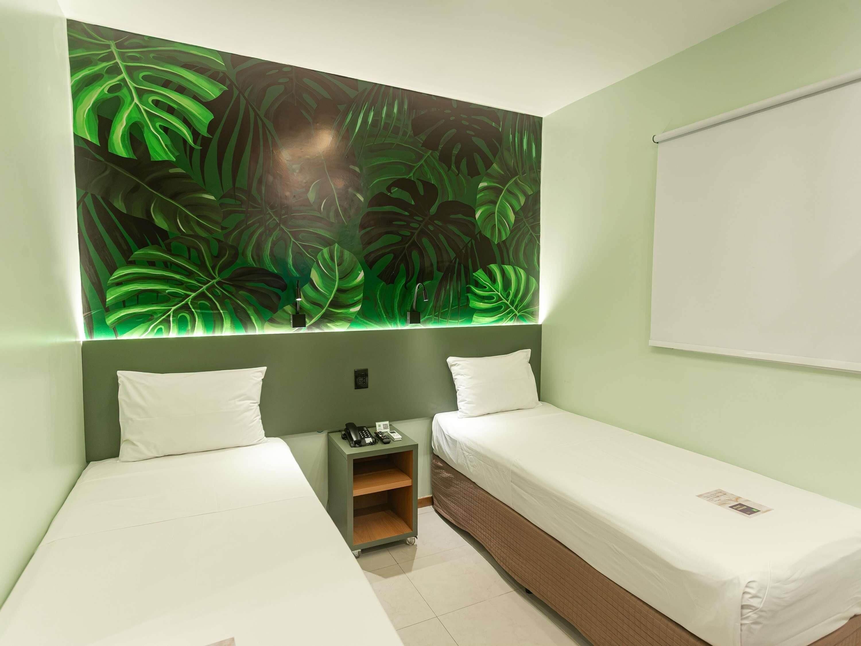 A hotel room with two single beds, a small bedside table with a lamp, and a large wall mural of green leaves.
