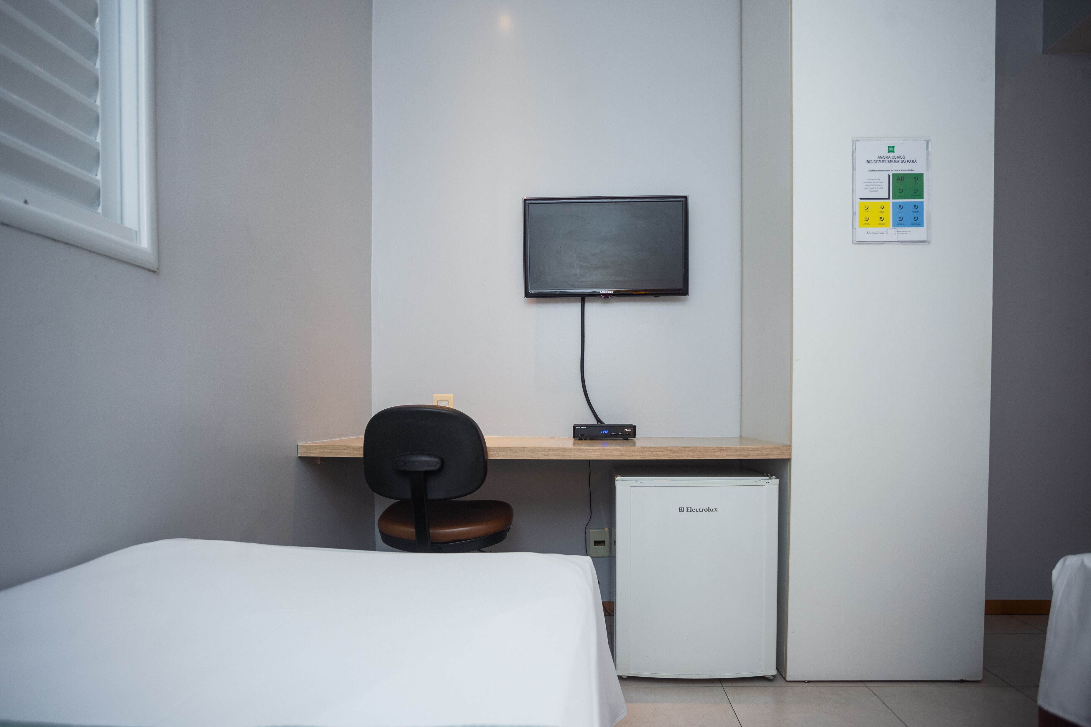 Habitación de hotel con cama, escritorio, monitor de computadora, silla y un refrigerador pequeño.