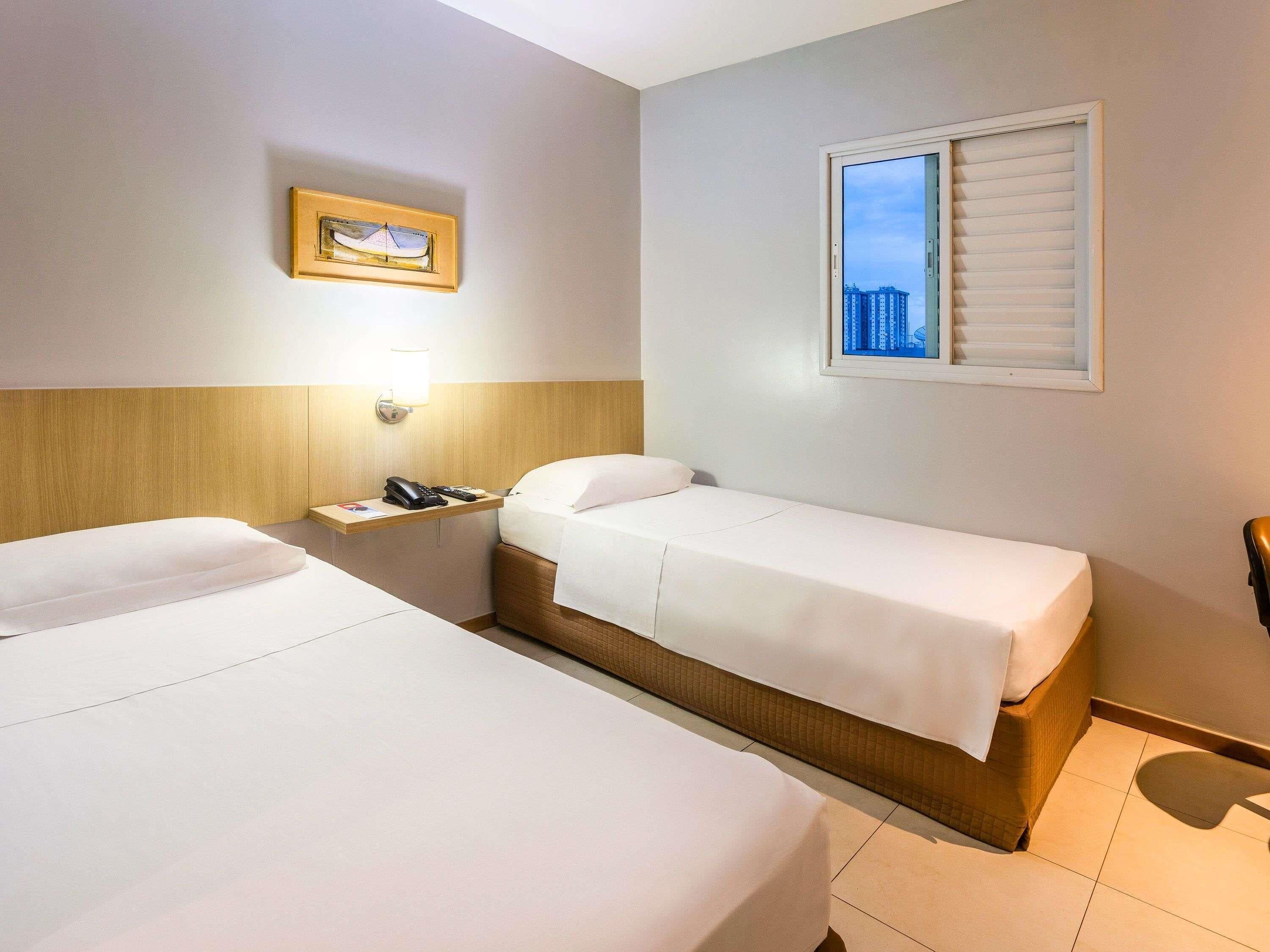 Habitación de hotel con dos camas individuales, una mesita de noche con lámpara, un cuadro en la pared y una ventana con persianas.