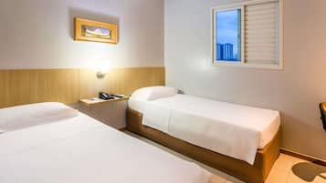 Habitación de hotel con dos camas individuales, una mesita de noche con lámpara, un cuadro en la pared y una ventana con persianas.