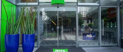 The entrance of a building with a glass facade, a green sign reading "ibis Styles", and two large blue planters.