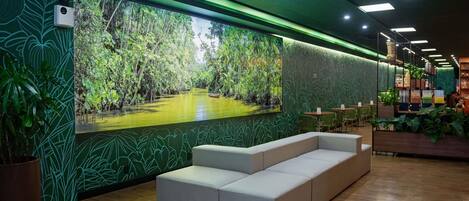 Un interior moderno con un amplio mural de un paisaje tropical, zona de estar y una sección con estanterías y plantas.