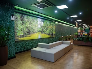 Un interior moderno con un amplio mural de un paisaje tropical, zona de estar y una sección con estanterías y plantas.