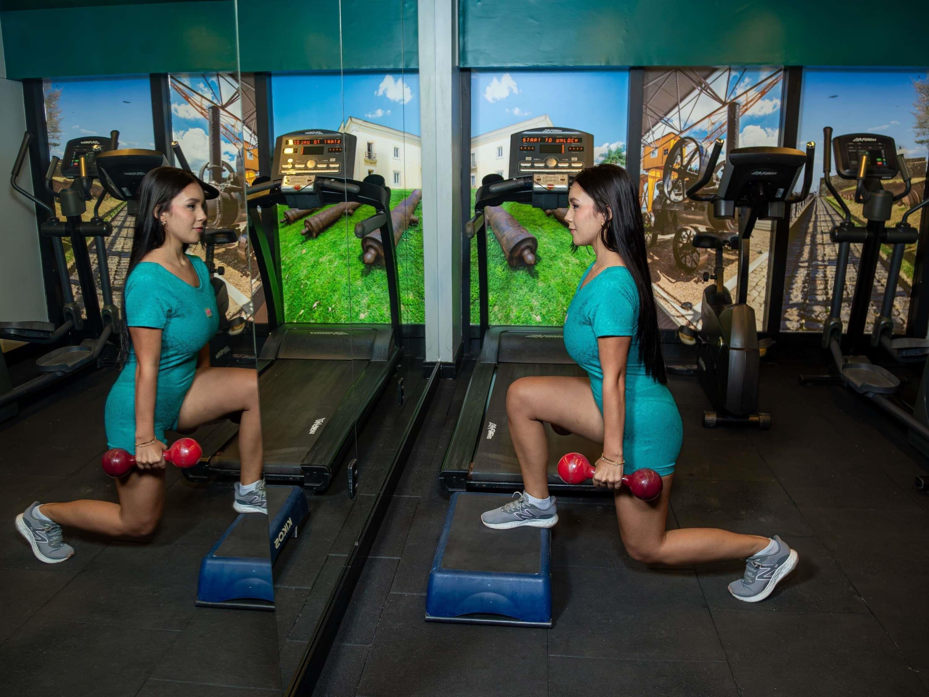 Dos personas realizando un ejercicio de sentadilla con mancuernas en un gimnasio con cintas de correr y equipo de ejercicio.