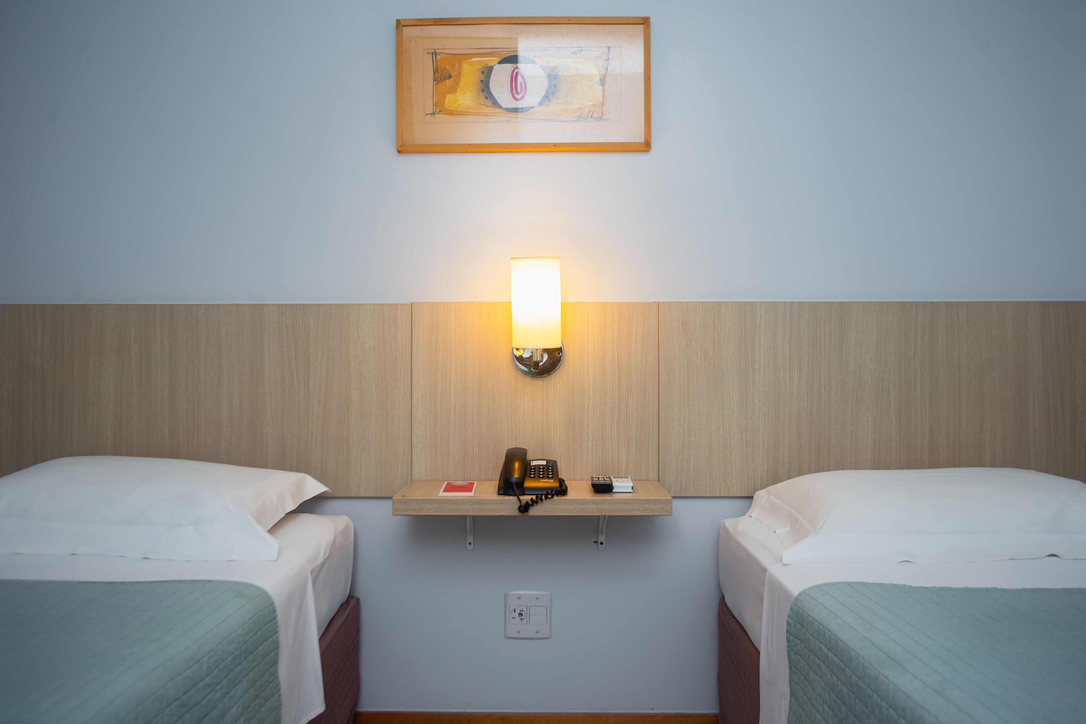 Una habitación de hotel con dos camas individuales, una mesita de noche con lámpara, teléfono y un cuadro en la pared.