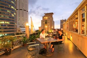 Una terraza en la azotea con mesas y sillas, plantas en macetas y vistas de edificios circundantes.