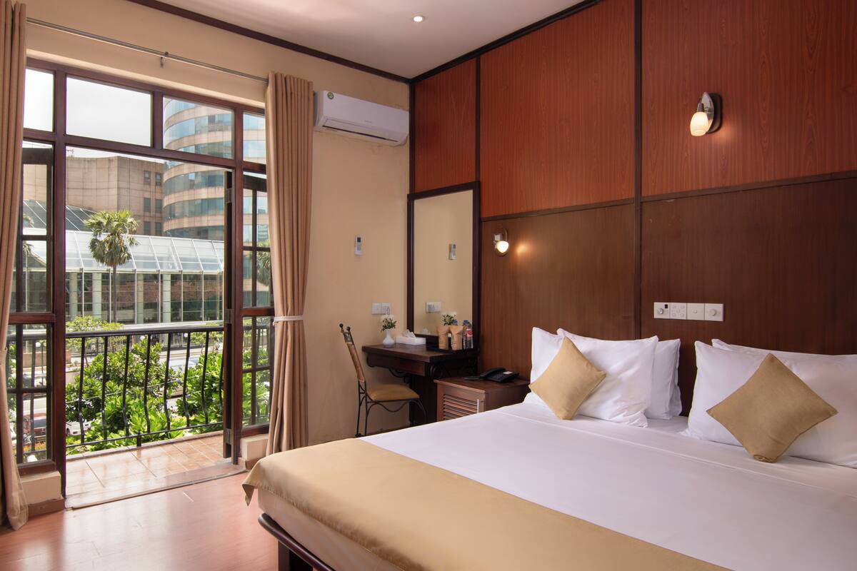 A hotel room with a large bed, a desk with a chair, a window with curtains, and a view of greenery outside.