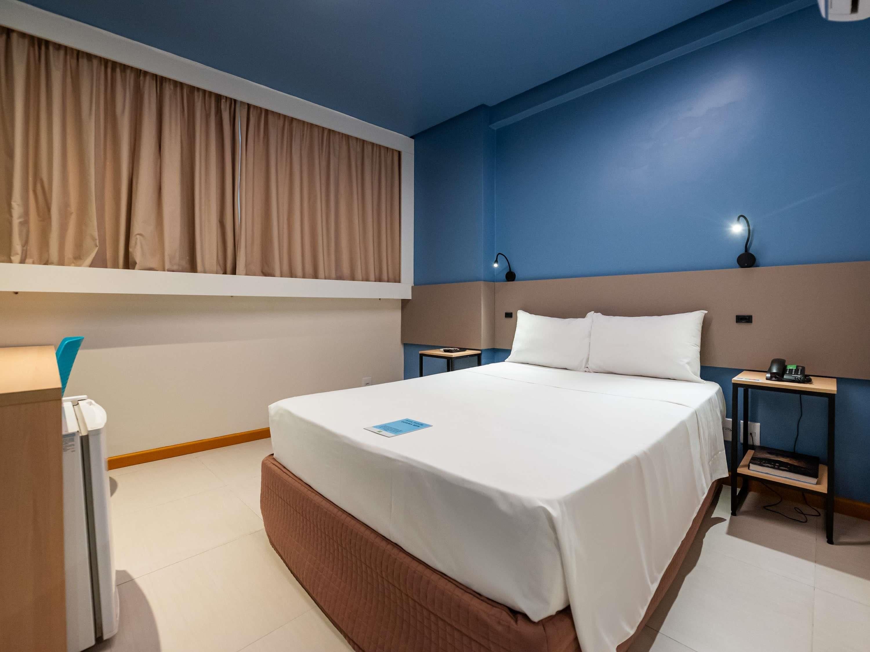 Una habitación de hotel con una cama grande, mesitas de noche, un escritorio con lámpara y una cortina.