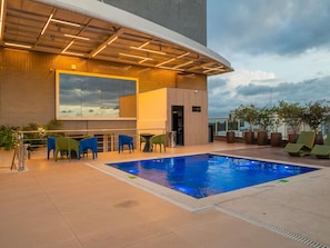 Un espace piscine aménagé sur le toit, comprenant une piscine, des chaises longues et un bâtiment moderne doté d’une façade en verre.
