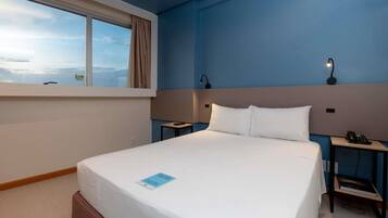 Una habitación de hotel con una cama grande, una mesita de noche con lámpara y otro buró con teléfono, además de un ventanal con cortinas.