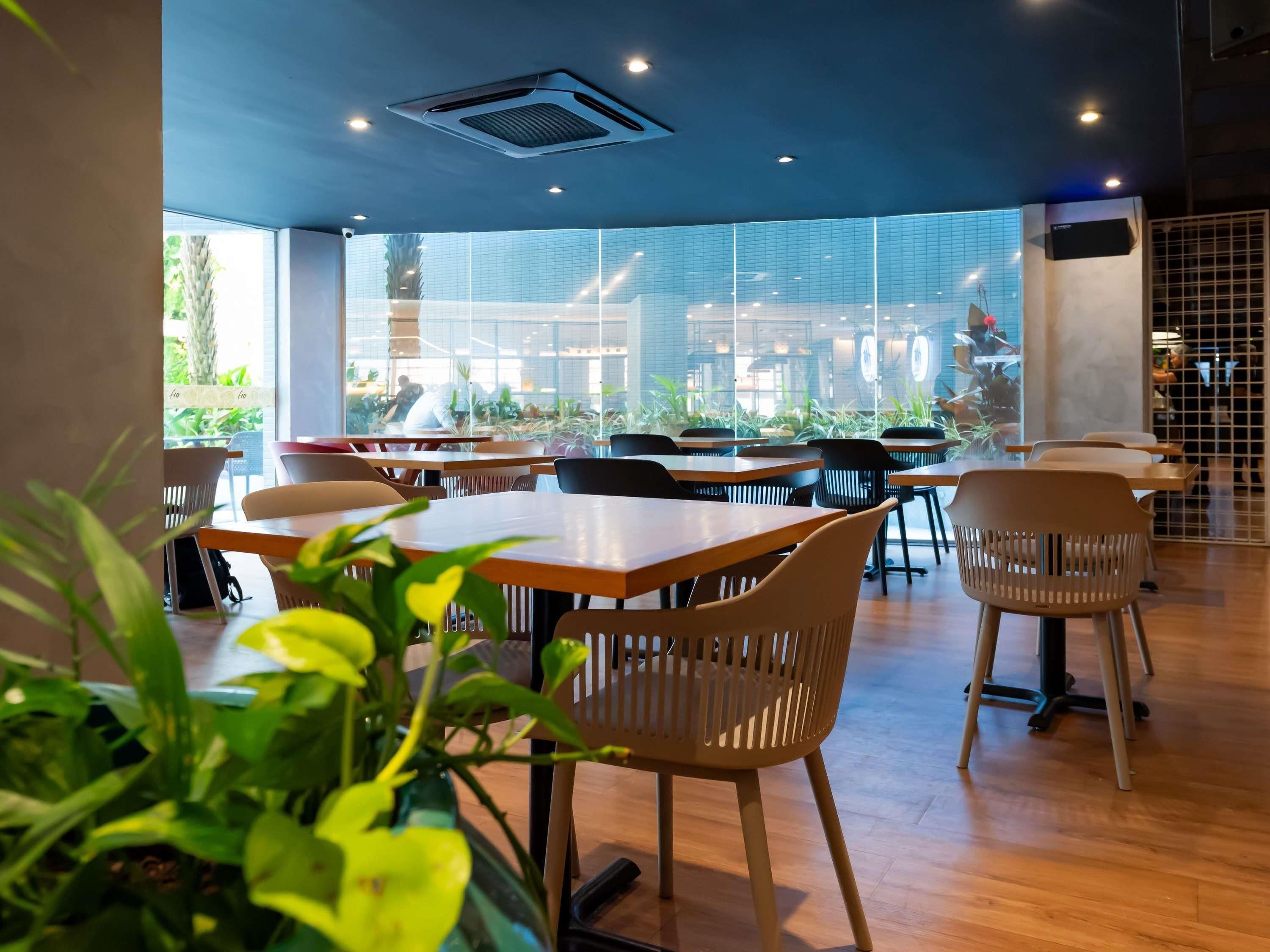 A modern cafe with wooden tables and chairs, large windows offering views of the outside, and a ceiling with recessed lighting.
