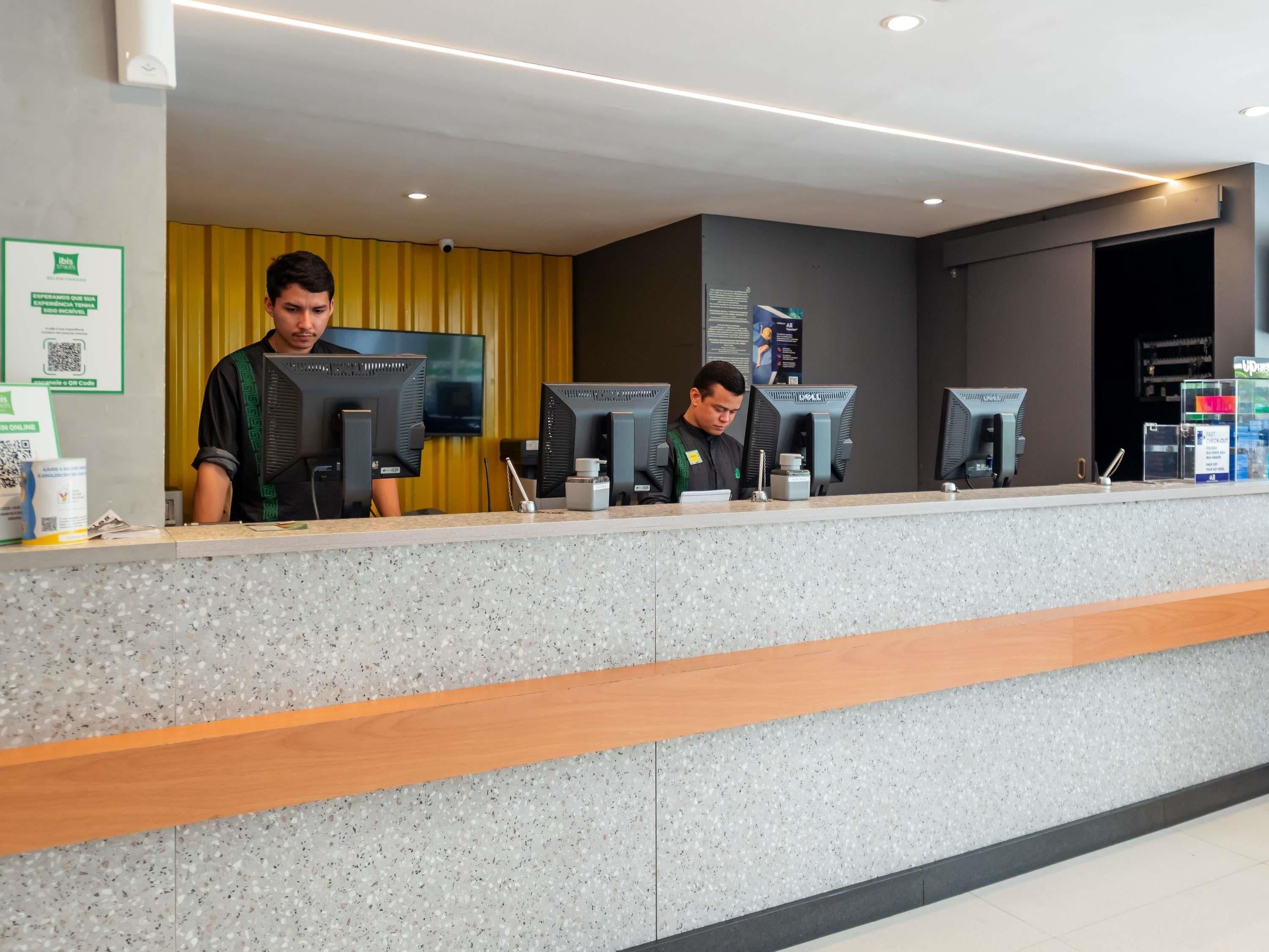 Un mostrador de recepción con dos empleados, una mesa de registro, una computadora y una pantalla montada en la pared.