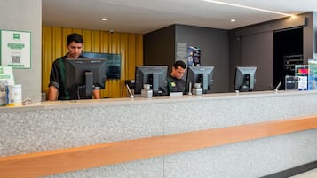 Un mostrador de recepción con dos empleados, una mesa de registro, una computadora y una pantalla montada en la pared.