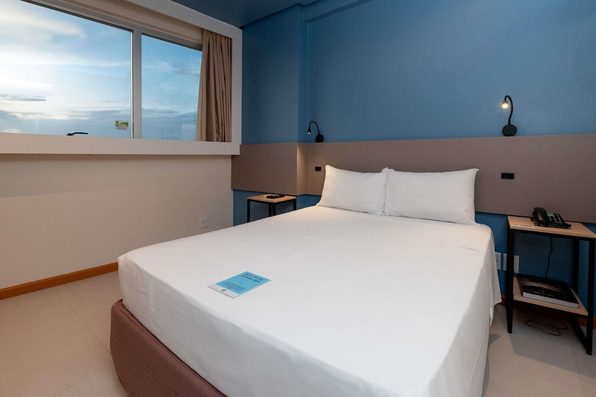 Una habitación de hotel con una cama grande, una mesita de noche con lámpara y otro buró con teléfono, además de un ventanal con cortinas.