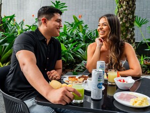 Deux personnes sont assises à une table à l’extérieur, devant de la nourriture et des boissons, et sont entourées de verdure.