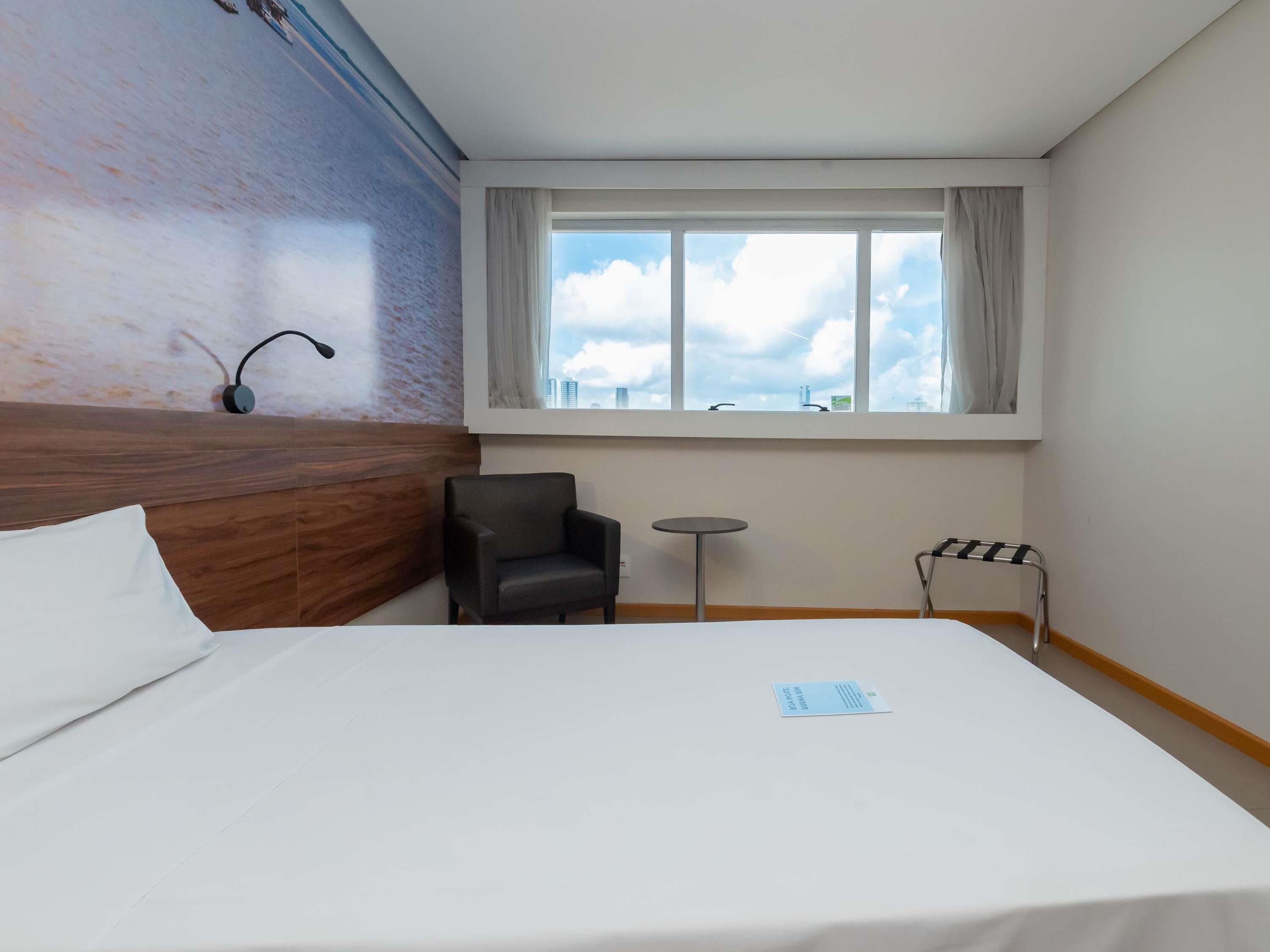A hotel room with a large bed, a small round table, a chair, a window with a view of the city, and a wall with a textured pattern.