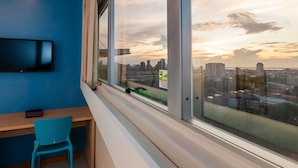 Een kamer met een groot raam met uitzicht op een stadsbeeld bij zonsondergang, een bureau met stoel, en een aan de muur bevestigde televisie.