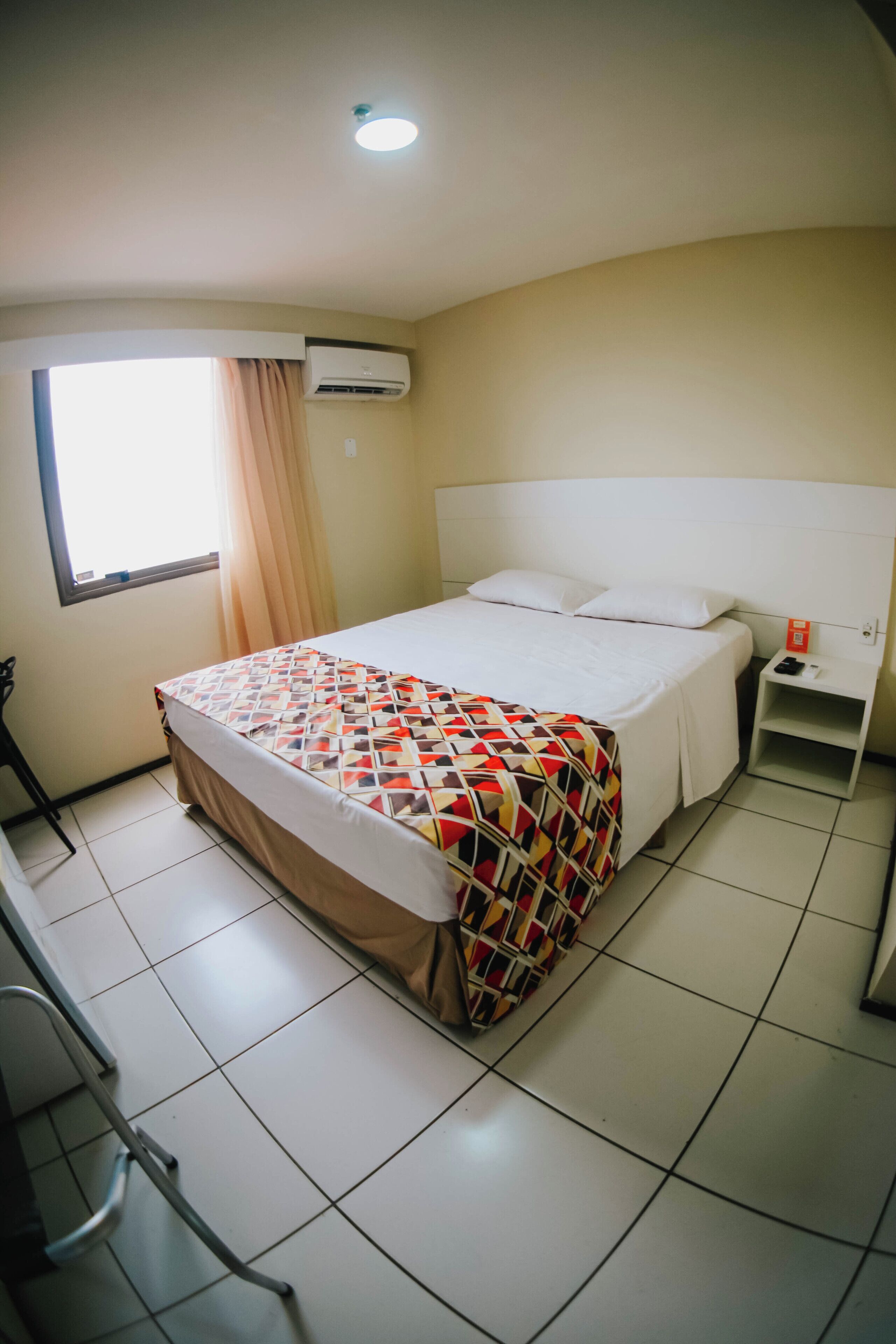 A hotel room with a bed, a nightstand, a window with curtains, and an air conditioning unit.