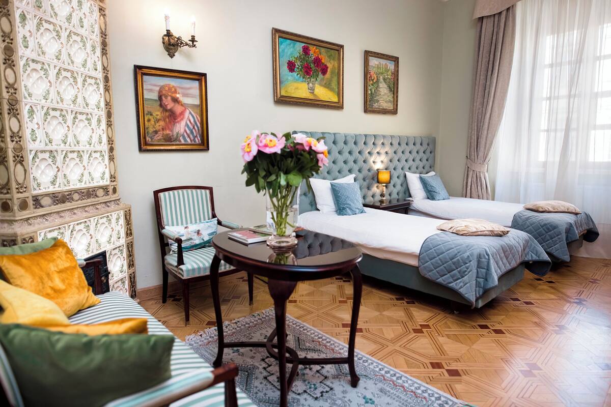Hotellihuone, jossa on napitettulla päätyosalla varustettu sänky, pieni pyöreä pöytä maljakko täynnä kukkia, raidallinen nojatuoli ja kuviollinen matto.