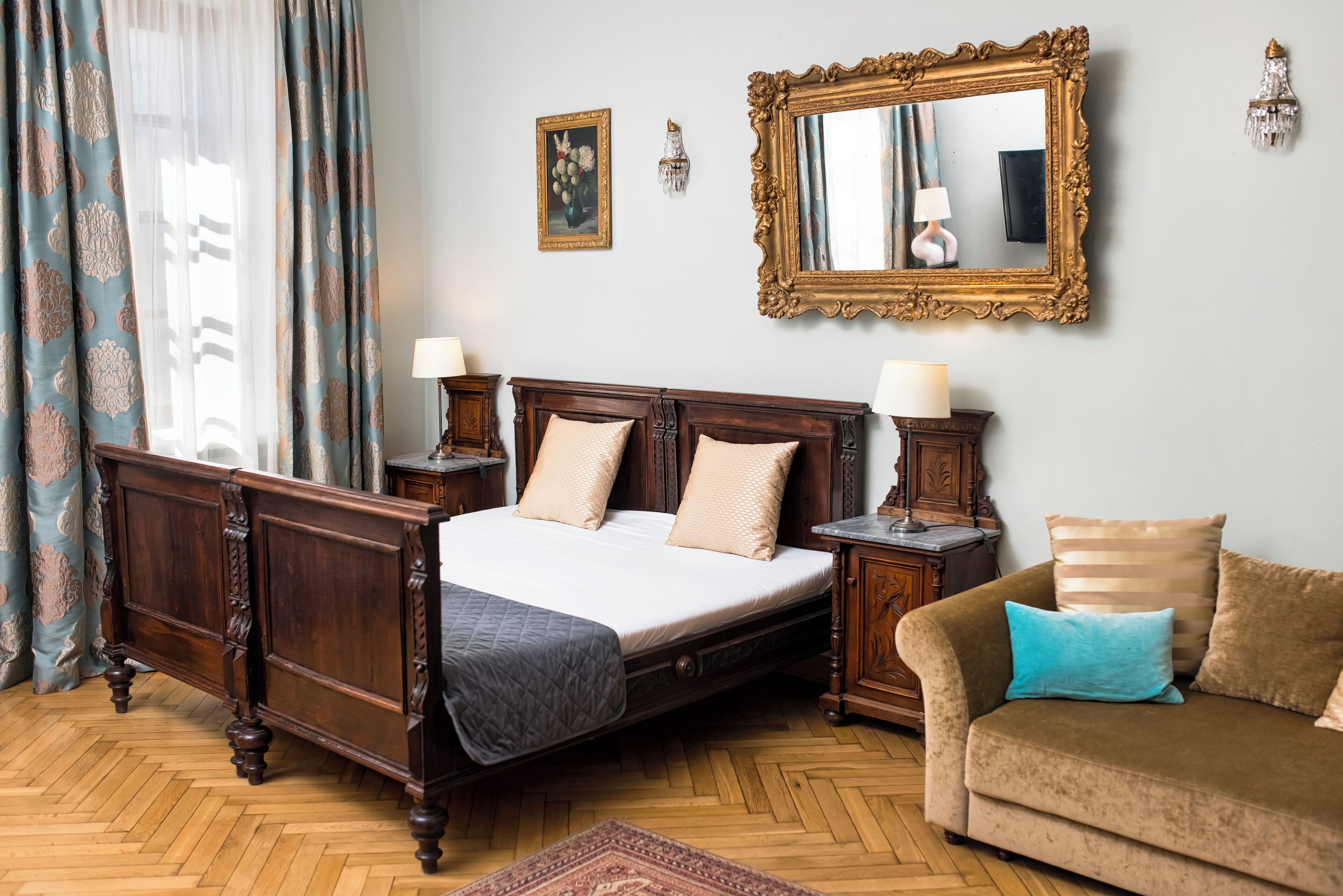 Un dormitorio con una cama con dosel de madera, dos mesitas de noche con lámparas, un sofá con cojines, un espejo grande y un aplique de pared.