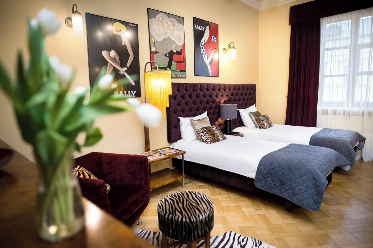 Hotellihuone, jossa on kaksi sänkyä, punainen nojatuoli, seeprankuvioinen jalkarahi, puinen sivupöytä, jossa on lamppu, ja julisteita seinällä.