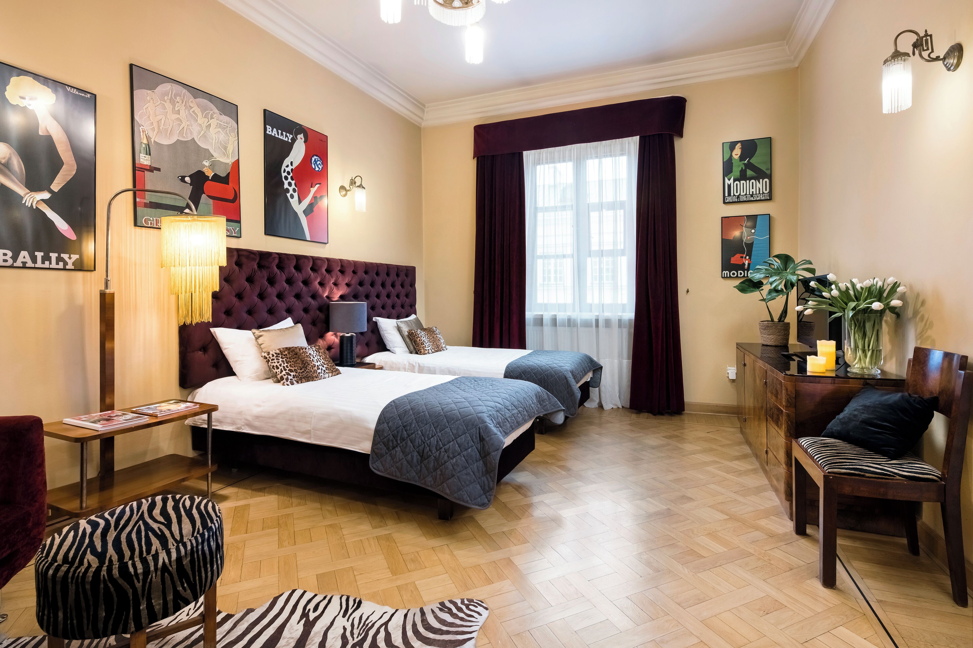 Une chambre d’hôtel avec deux lits, une tête de lit capitonnée, un plancher en bois et des œuvres d’art accrochées aux murs.