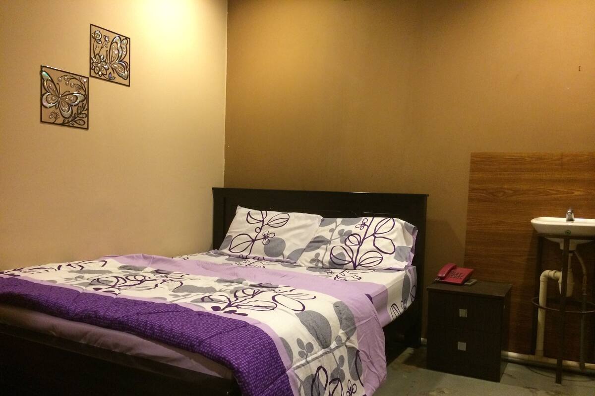 A bedroom with a bed, a nightstand, a wall-mounted phone, and a bathroom with a sink.