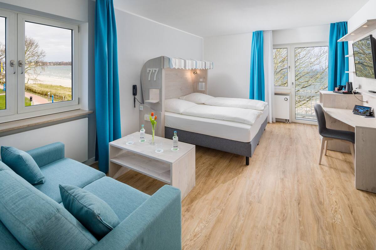 A modern hotel room with a bed, a desk, a sofa, and a view of the sea.