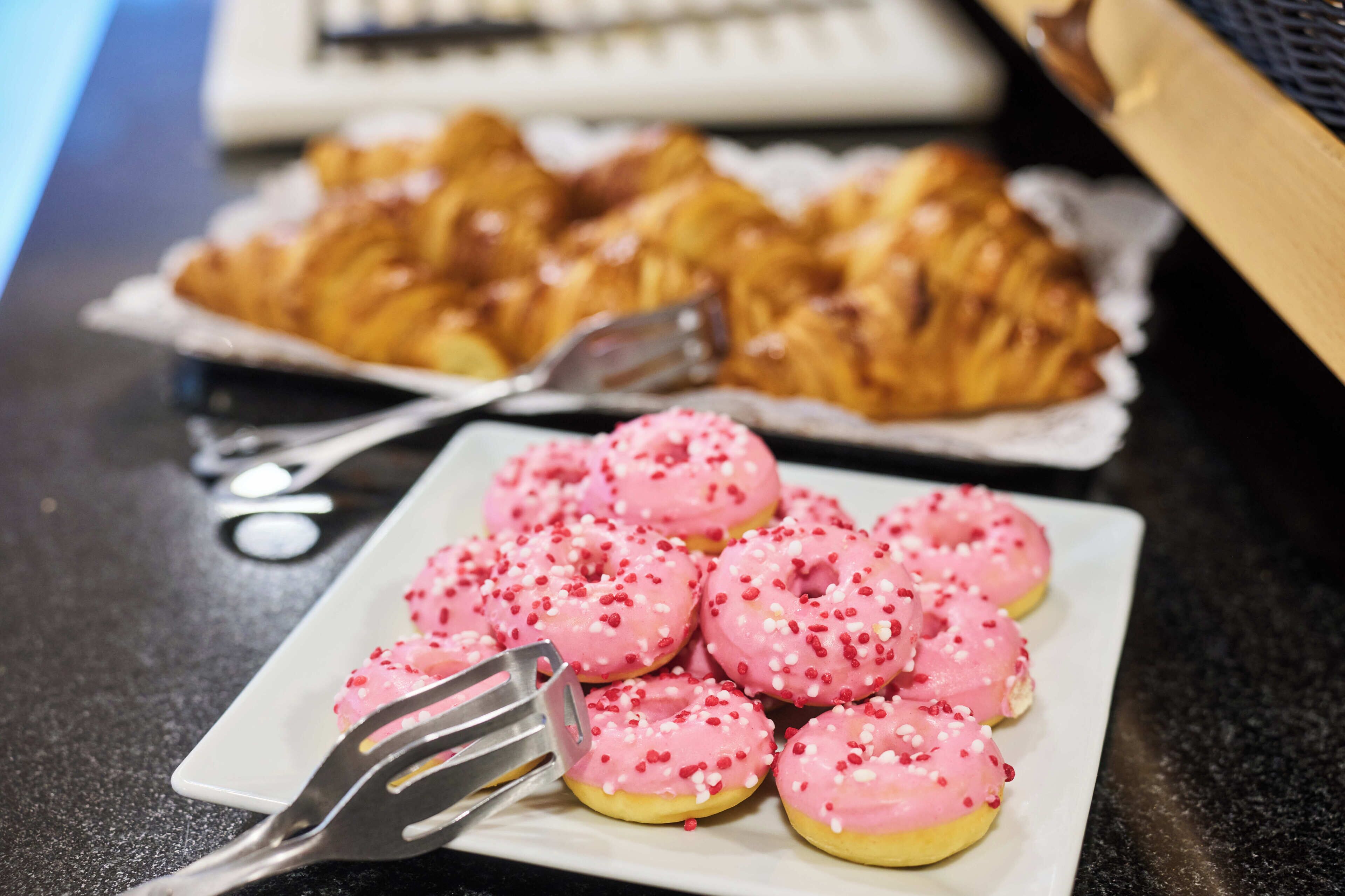 Un plato de donas rosas cubiertas de glaseado y chispas, una bandeja de cruasanes y un par de cubiertos sobre una encimera oscura.