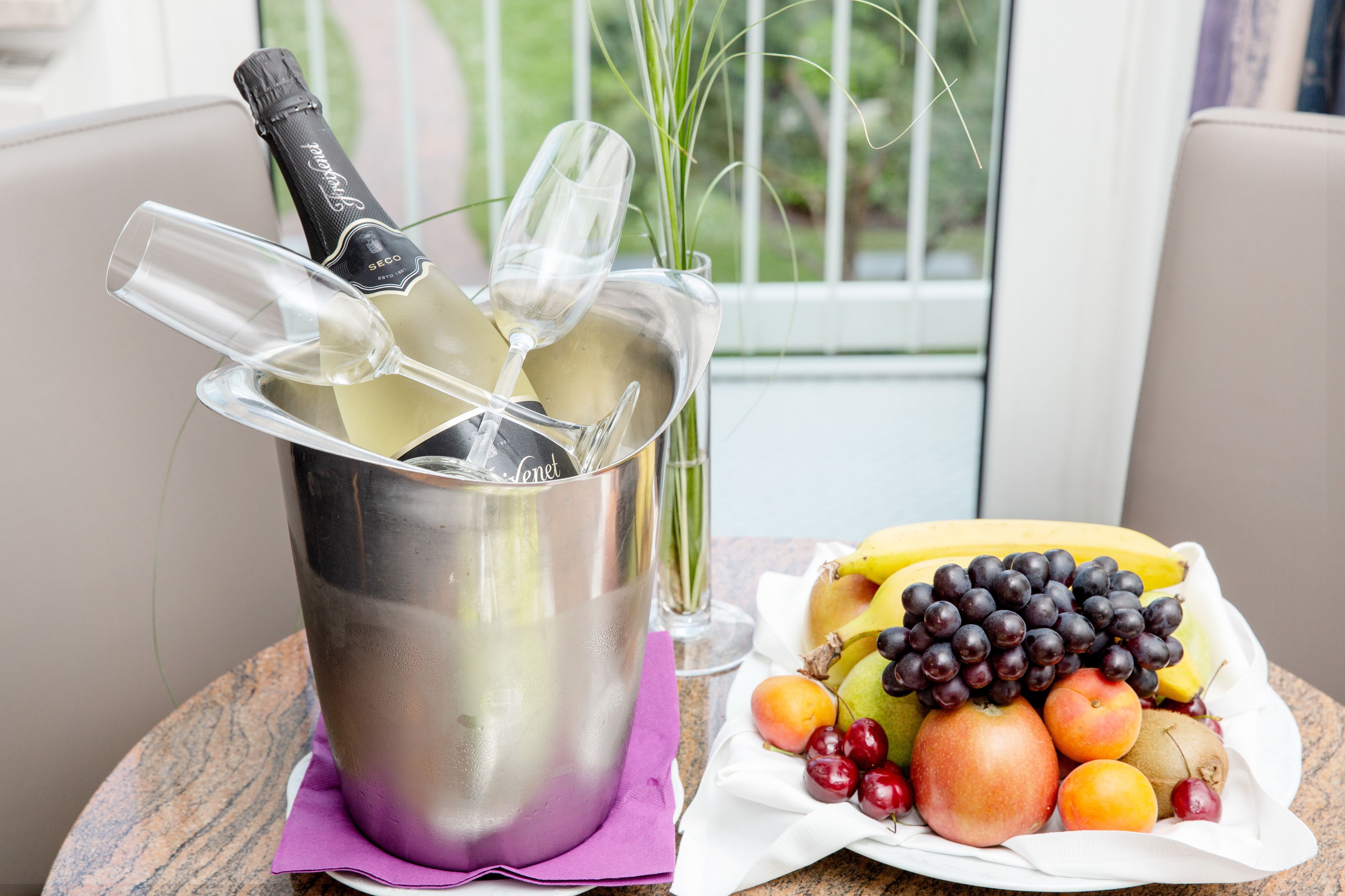 Un balde de metal con una botella y vasos, un cuenco de fruta, y vista al exterior por la ventana.