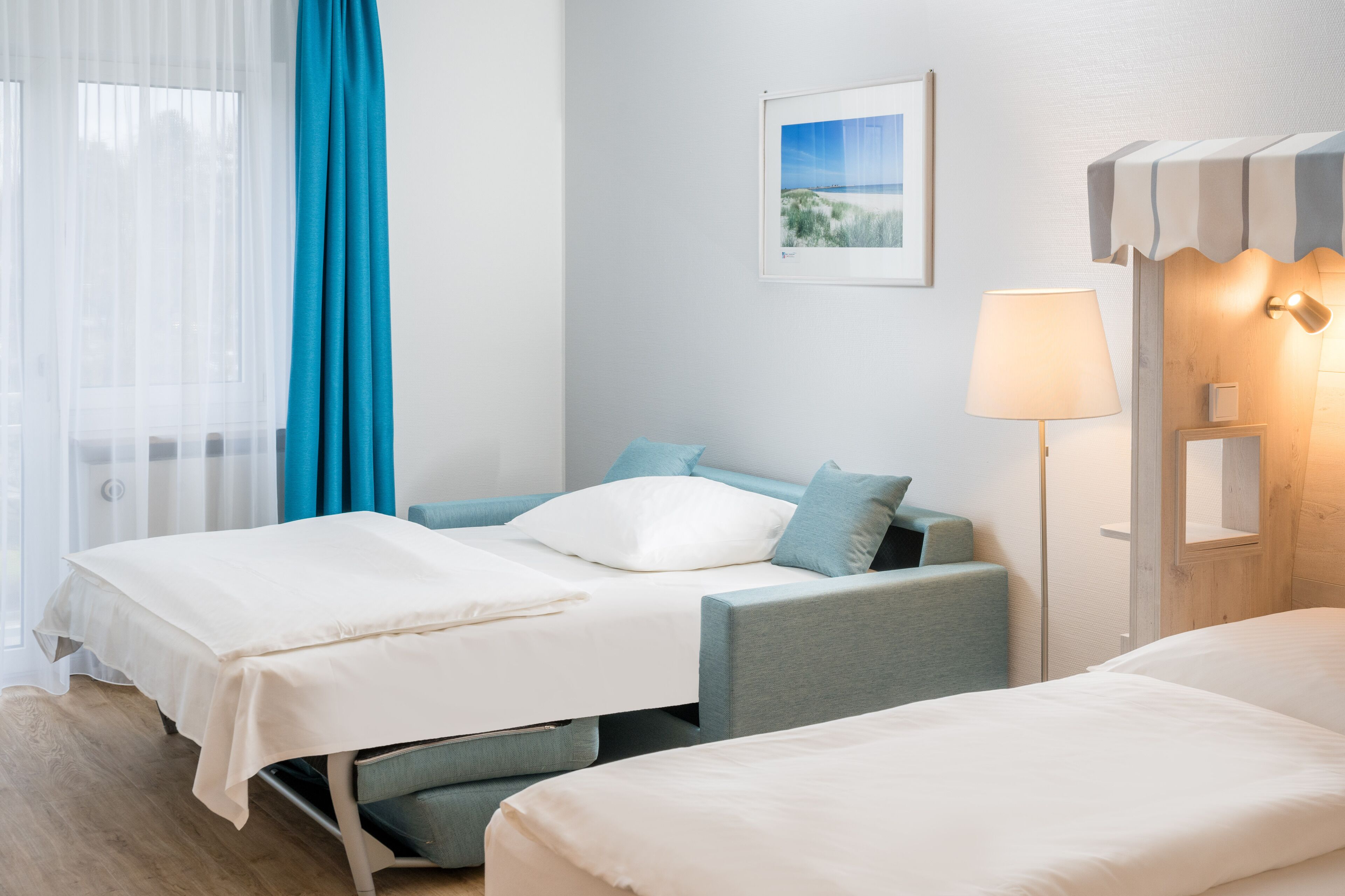 Habitación de hotel con dos camas, un ventanal con cortinas translúcidas, una lámpara de pared y un cuadro enmarcado.