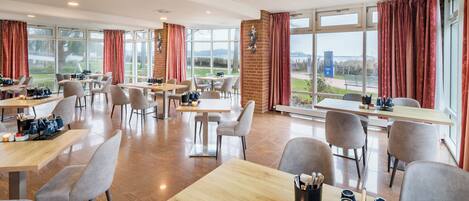 A modern dining area with tables and chairs, large windows with curtains, and a view of the outdoors.