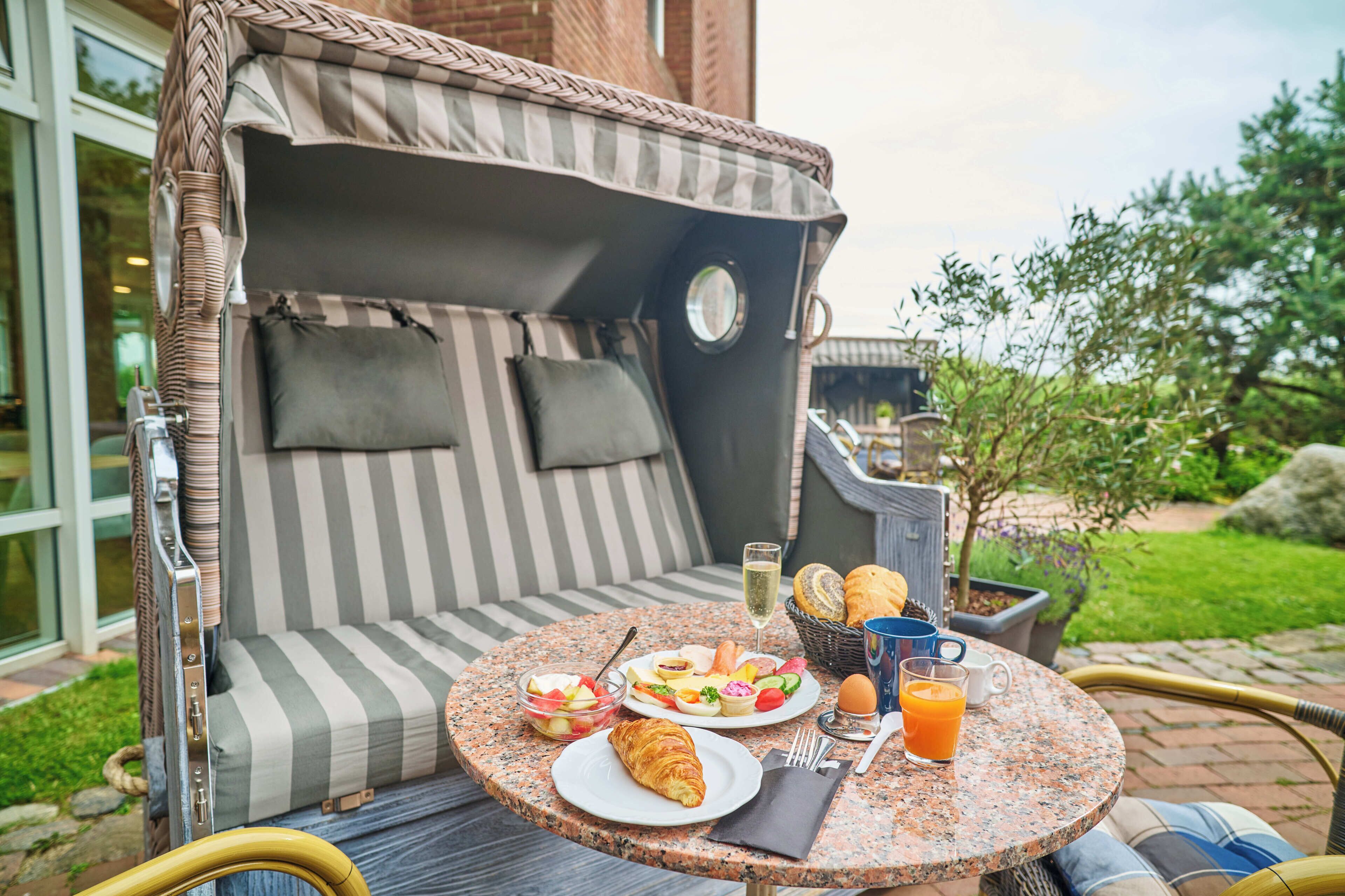 A wicker outdoor seating area with a table set for breakfast, including croissants, fruit, and beverages.