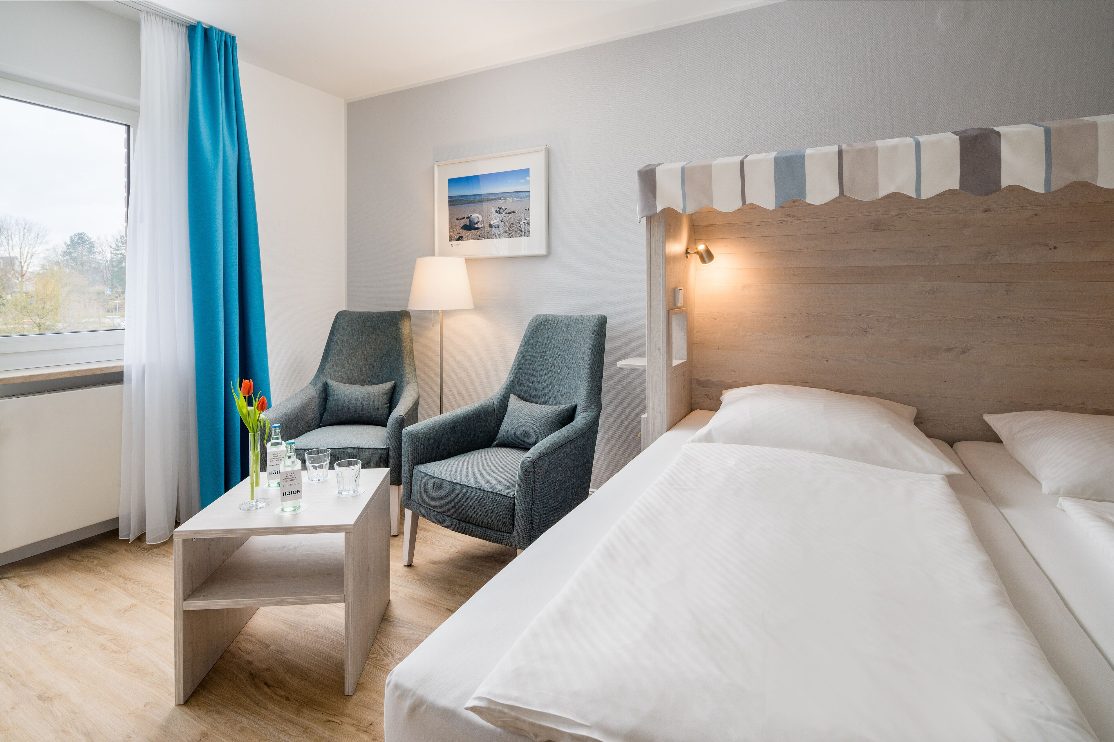A hotel room with a bed, two armchairs, a small table with a vase of flowers, a lamp, and a framed picture on the wall.