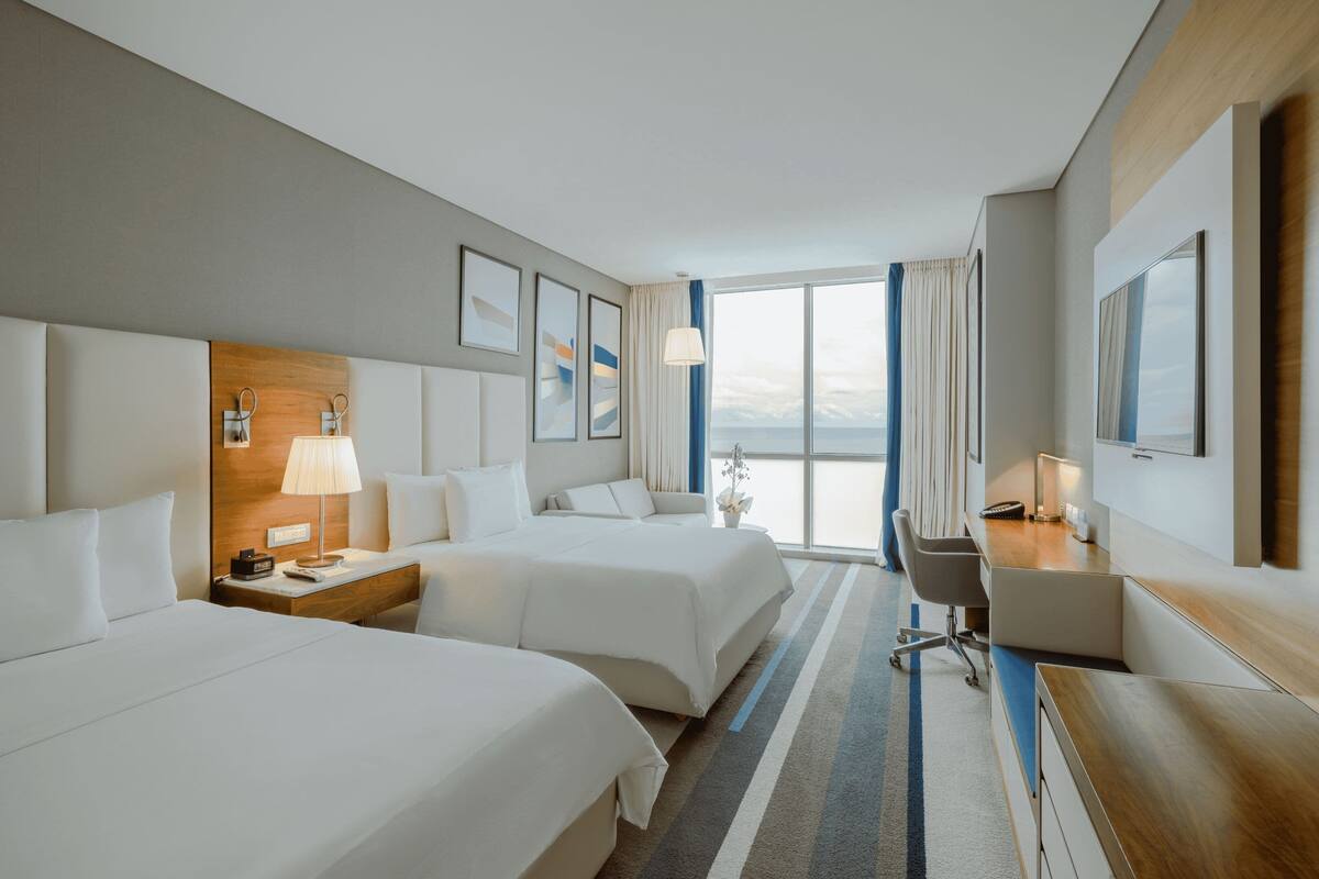 Een hotelkamer met twee bedden, een bureau, een stoel, een lamp en uitzicht op zee.