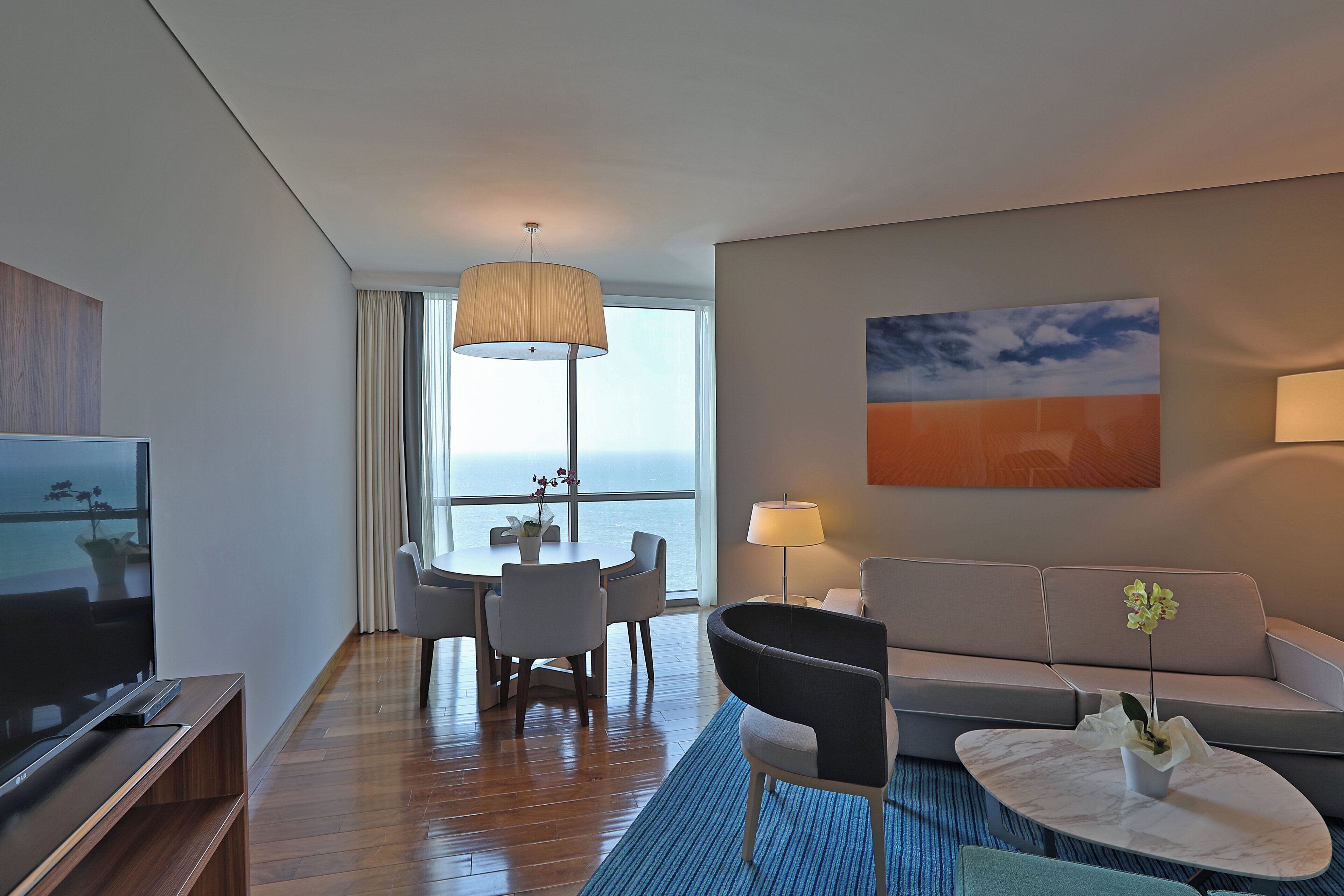 A modern hotel room with a flat-screen TV, a sofa, a dining table with chairs, a coffee table, and a large window with a view of the sea.