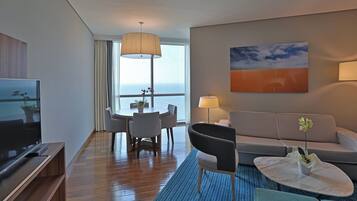 A modern hotel room with a flat-screen TV, a sofa, a dining table with chairs, a coffee table, and a large window with a view of the sea.