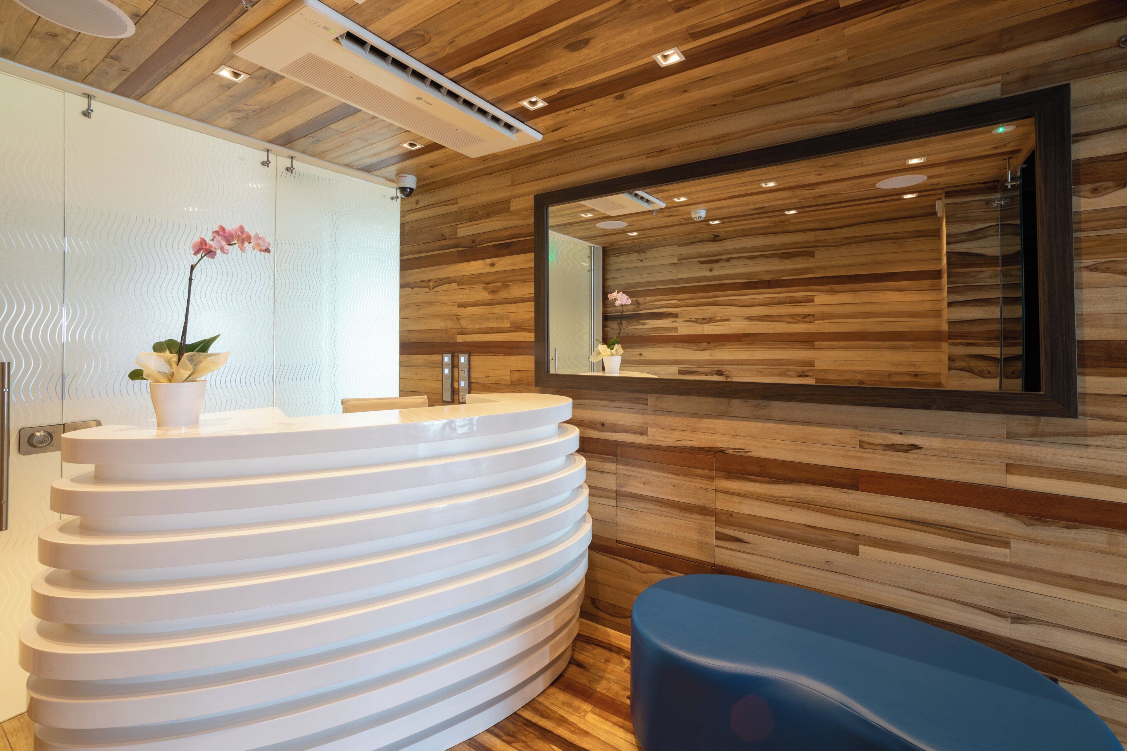 A modern bathroom with a white, curved vanity, a wooden wall panel, a large mirror, and a blue bench.