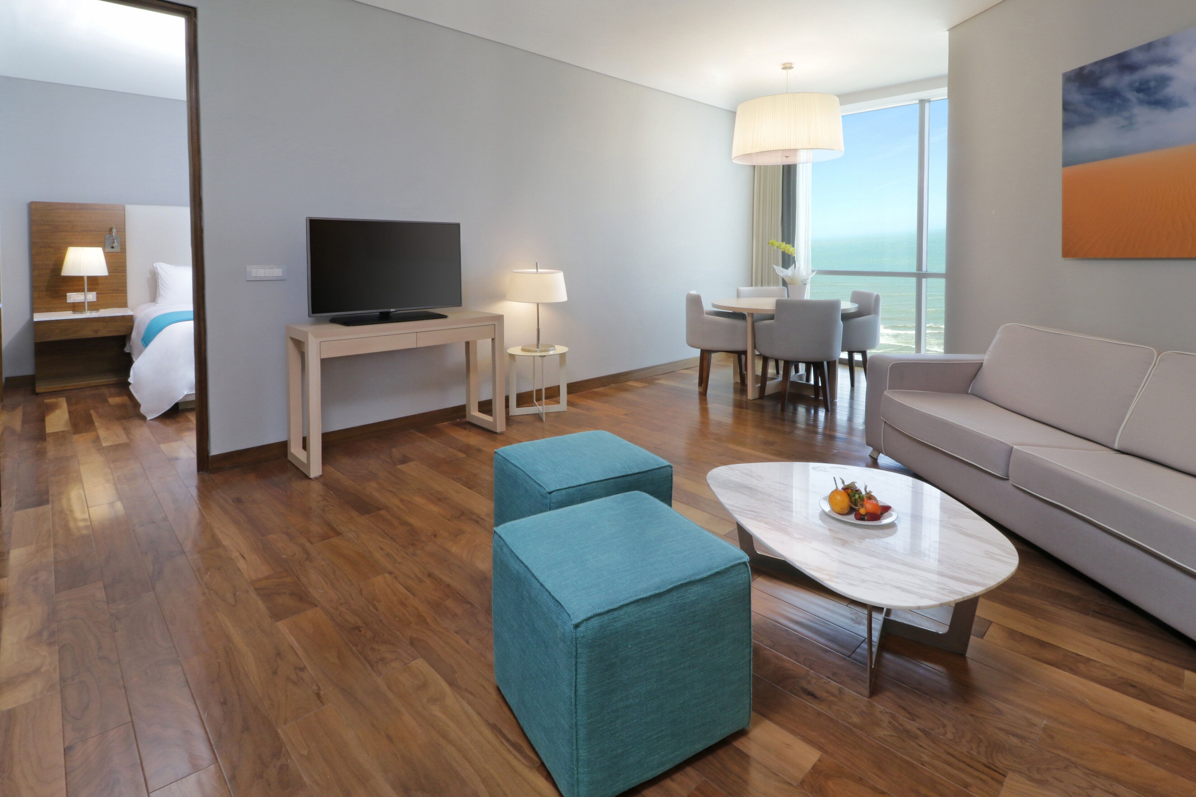 Habitación de hotel moderna con piso de madera, televisor de pantalla plana, sofá, mesa de comedor con sillas y un ventanal con vistas.