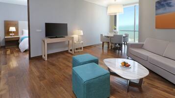 Habitación de hotel moderna con piso de madera, televisor de pantalla plana, sofá, mesa de comedor con sillas y un ventanal con vistas.