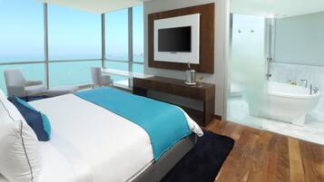 A modern hotel room with a large bed, a TV, a glass-enclosed shower, and a view of the sea.