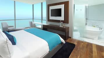 Habitación de hotel moderna con una cama grande, televisor, ducha con paredes de vidrio y vista al océano.
