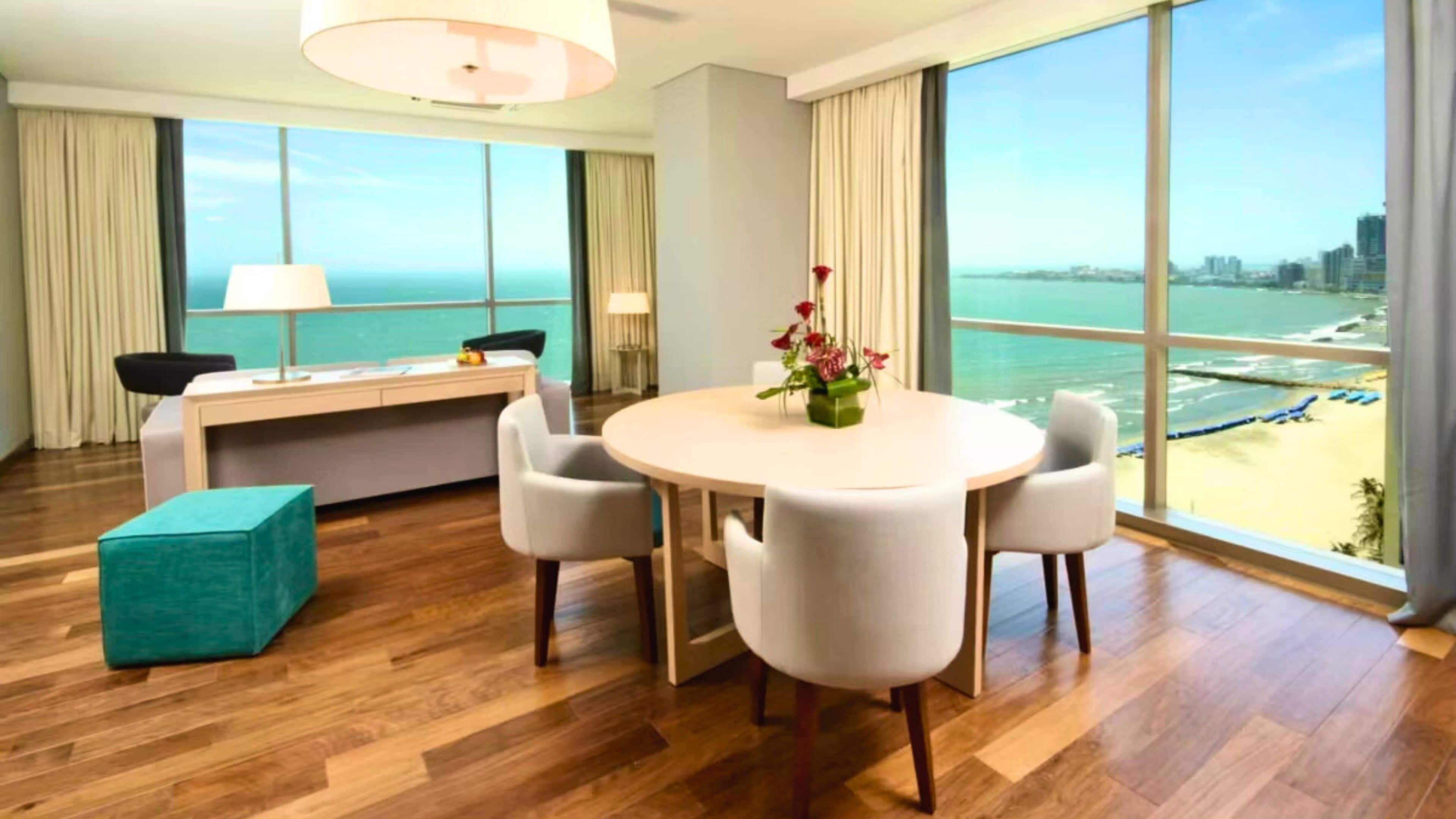 Habitación de hotel con piso de madera, una mesa redonda de comedor con sillas, un escritorio y una silla, un ventanal con vista a la playa y al océano, y un otoman de color turquesa.