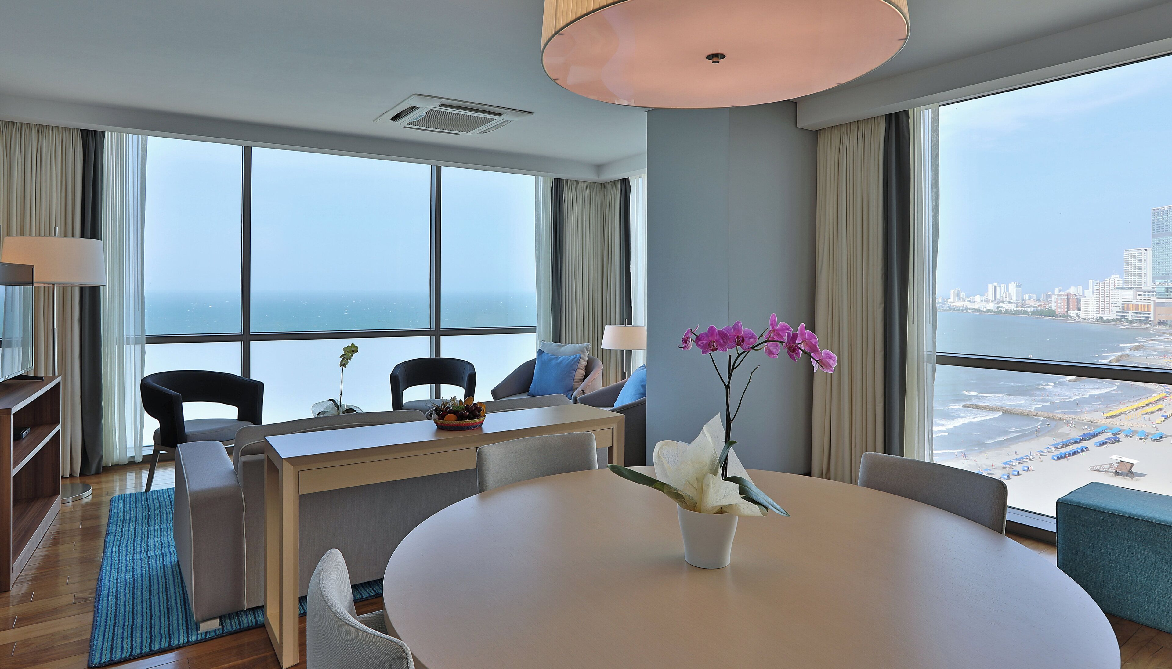 A modern hotel room with a large window, a round dining table, and a view of the city and beach.