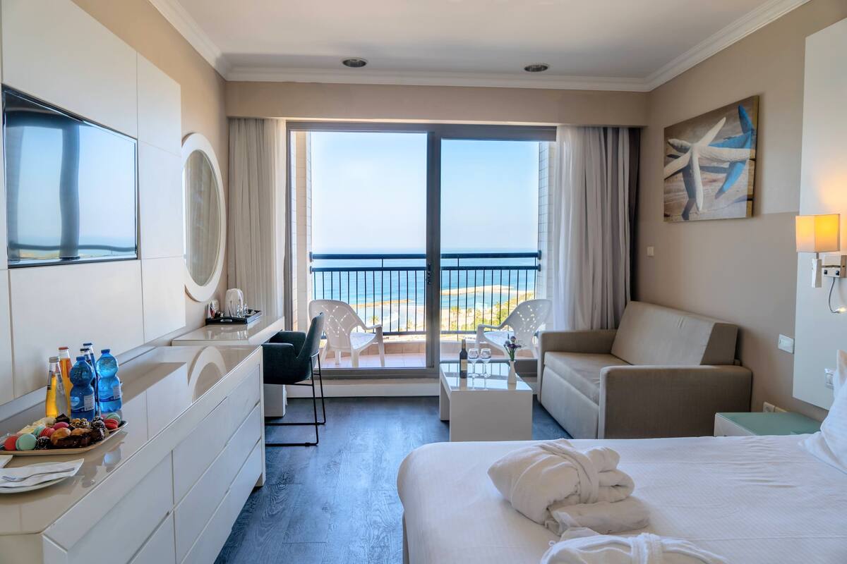 Habitación de hotel con cama, escritorio, sillón, sofá, balcón con vistas a la playa y un ventanal grande.